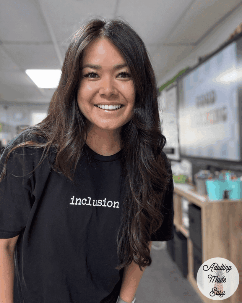 A woman with dark long hair standing and smiling in her classroom.