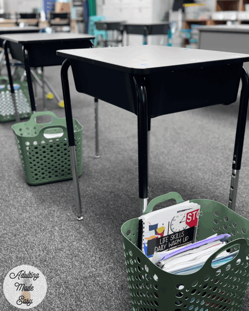 Student desks in a classroom with a green basket next to the desks containing daily warm up booklets and other work materials.